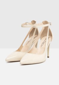 Décolleté in vernice beige con punta affusolata, cinturino alla caviglia e tacco a stiletto. Texture liscia e design minimalista.
