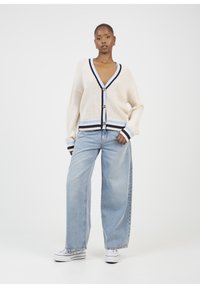Cardigan crema con un profondo scollo a V, accenti a righe blu scuro e blu chiaro, grandi bottoni, indossato sopra jeans a gamba larga blu chiaro.