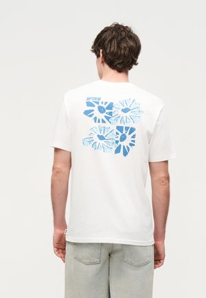 Mann iført hvit t-skjorte med fire blå abstrakte blomstermønstre på ryggen, kombinert med lysegrå denimshorts.