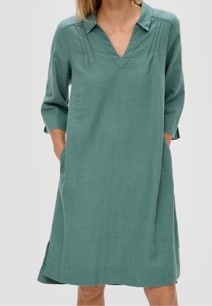 Femme portant une robe verte ample avec des manches trois-quarts, un col en V et les mains dans les poches latérales.