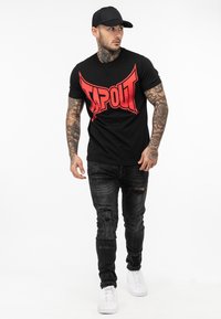 Sort kortærmet t-shirt med stort rødt "TAPOUT" logo, parret med slidte sorte jeans og hvide sneakers. Mand iført en sort cap.