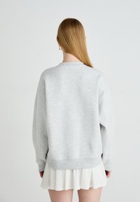 Pessoa com cabelo louro comprido, vestindo um sweat-shirt cinza claro e uma saia plissada branca, de costas, contra um fundo claro e liso.