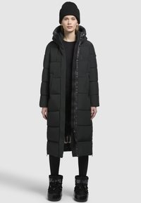 Schwarze Puffermantel mit langem Design, gesteppter Textur, Frontreißverschluss und Kapuze. Getragen über einem schwarzen Oberteil; kombiniert mit schwarzen Stiefeln und Leggings.