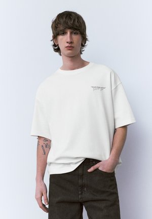 Junger Mann mit Ohrringen und Tattoos, der ein weißes übergroßes T-Shirt mit dem Text "Streets keep secrets" und schwarze Hosen trägt, steht vor einer weißen Wand.