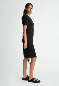 Black short-sleeved dress with a straight cut, side drawstrings for adjustment, worn with black slide sandals featuring gold buckles.