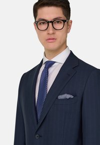 Junger Mann mit schwarzen runden Brillen, marineblauem Nadelstreifenanzug, weiß gestreiftem Hemd, blau gemusterter Krawatte und gemustertem Einstecktuch vor weißem Hintergrund.