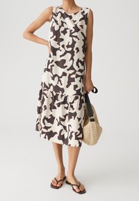 Femme portant une robe sans manches longueur genou avec un imprimé abstrait noir et blanc, tenant un sac tissé beige et portant des tongs noires.