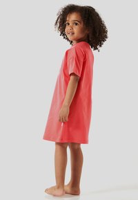 Kleid aus korallenfarbenem Baumwoll-T-Shirt mit kurzen Ärmeln, knielangem Design und seitlichen Schlitzen. Das Modell steht barfuß und blickt leicht nach links.