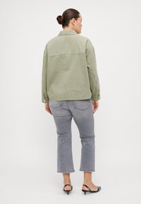 Olive green denim jacket with a collared neckline, snapped cuffs, and seam detailing, paired with gray cropped jeans and black heeled sandals.