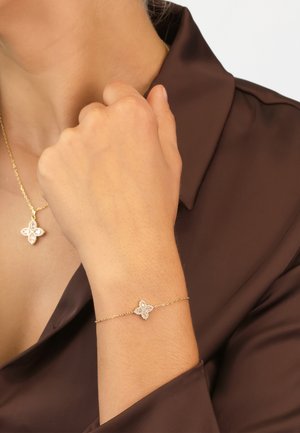 Gold bracelet with a floral charm featuring circular crystal accents, worn on a wrist, complemented by a matching pendant necklace.