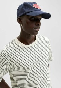 Person iført en navy kasket med rød logo og en hvid skjorte med tynde sorte vandrette striber, vendt let nedad og mod højre.