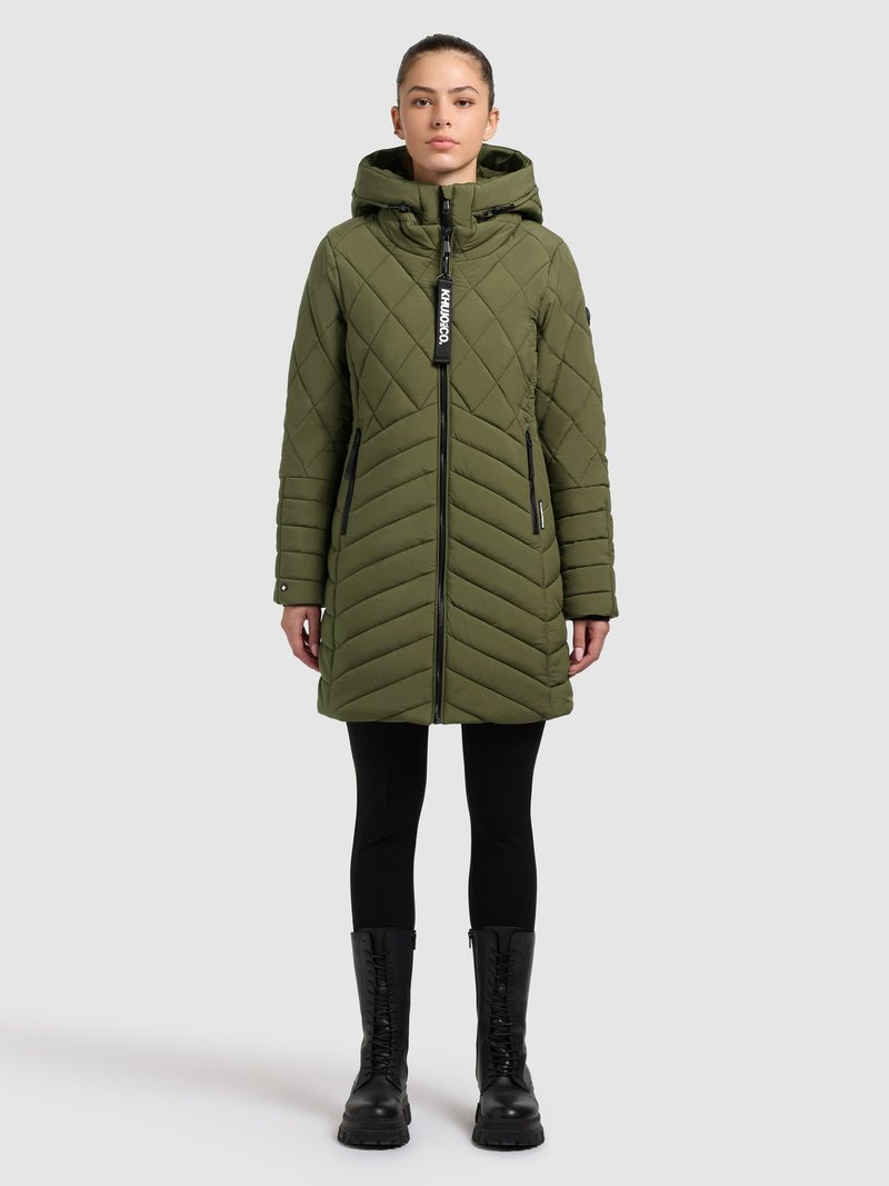 Olívazöld, steppelt pufi kabát kapucnival, cipzáras záródással, két oldalzsebbel és márkajelzéses címkével; fekete leggingsszel és fűzős csizmával párosítva.