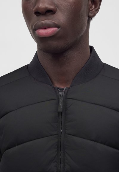 Gros plan d'une personne portant une veste noire matelassée avec une fermeture éclair étiquetée "CLOSED" et un col côtelé.