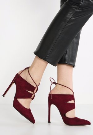 Vrouw die bordeauxrode suède hoge hakken met enkelbandjes draagt, gecombineerd met zwarte cropped leren broek, staand op een witte vloer.