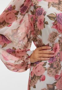 Main sur la taille portant un chemisier blanc transparent avec un motif floral rose et violet et des manches longues bouffantes.