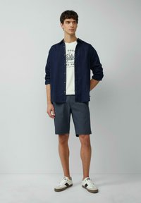 Marine linnen overhemd, wit graphic T-shirt, grijze shorts, witte sneakers met bruine accenten. Model staat tegen een effen achtergrond.