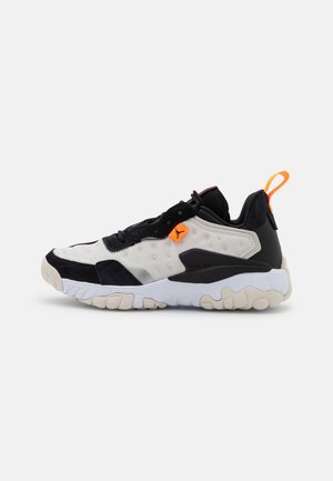 Baskets de sport noires, blanches et beiges avec semelle épaisse, languette de tirage orange au talon et petite étiquette orange avec un logo près des lacets.