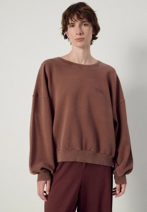 Persoon met kort bruin haar, gekleed in een losse bruine trui en bijpassende broek, staand tegen een effen lichte achtergrond.
