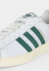 Λευκό sneaker Adidas Superstar με πράσινες ρίγες, λευκά κορδόνια και χρυσό κείμενο "Superstar" στο πλάι.