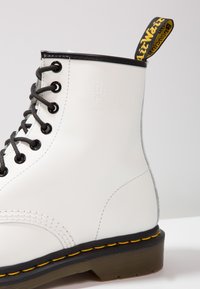 Botín de cuero blanco con cordones, con detalles en negro y cordones grises. Presenta un logo en relieve, costuras amarillas y una lengüeta en el talón.