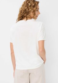 Hvid t-shirt med korte ærmer og rund hals, lavet af blødt materiale, båret med lyse beige bukser. Bagsiden viser lette syninger.