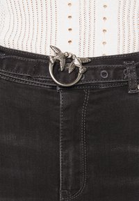 Svarta jeans med en silverfärgad ringb älte spänne med två intrikat designade fåglar. Texturerad tyg och figurnära passform är synlig.