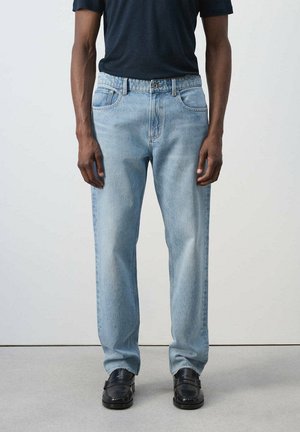 Persona que lleva vaqueros azul claro de pierna recta, zapatos negros y una camisa oscura de manga corta, de pie contra un fondo liso.