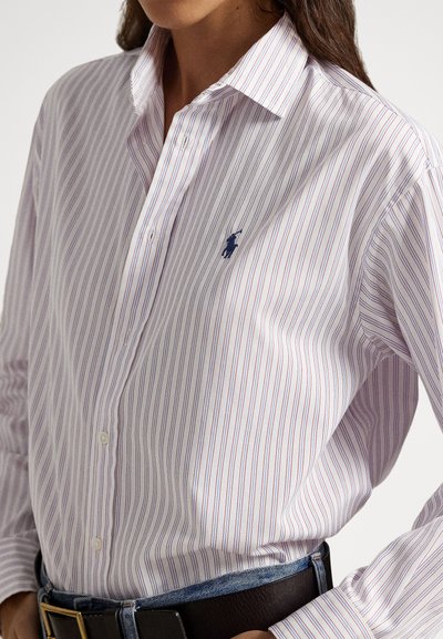 Sieviete valkā baltu kreklu ar plānām sarkanām un zilām vertikālām svītrām, ielikts džinsās ar melnu jostu.