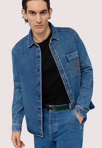 Koszula dżinsowa w średnim odcieniu niebieskiego, zapinana na guziki, z kieszenią na piersi. Posiada kołnierzyk i kontrastowe przeszycia. Nosi się ją na czarną koszulkę.