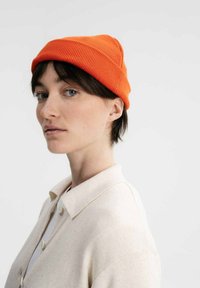 Oranje geribde beanie, gedragen met een omgeslagen rand. Model draagt een lichtbeige trui met een kraag. Neutrale achtergrond.