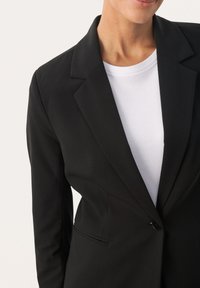 Blazer noir au design structuré, avec des revers nets et une fermeture à un bouton. Porté par-dessus un t-shirt blanc à col rond.