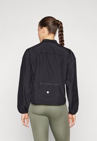 Zwarte lichte jas met een ritszak op de rug, geribbelde textuur, hoge kraag en elastische manchetten, gecombineerd met olijfgroene leggings.