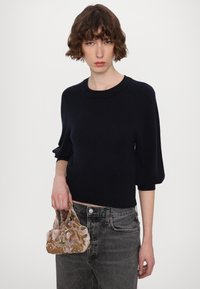 Schwarzer Strickpullover mit Rundhalsausschnitt und Puffärmeln, kombiniert mit dunkelgrauen Jeans. Hält eine kleine verzierte Samttasche mit floralen Mustern.