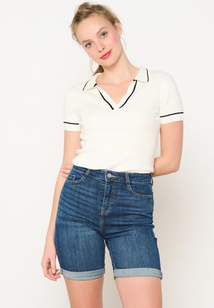 Bela polo majica s kratkimi rokavi in črnimi dodatki, v kombinaciji s temno denim kratskimi hlačami, zvijanimi pri robu. Prijeten kroj, gladka tekstura in klasičen dizajn.