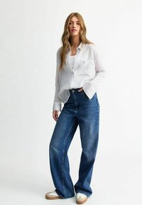 Vit och grå randig skjorta med knappar, avslappnad passform, i kombination med högmidjade blå jeans. Modellen bär vita sneakers med bruna detaljer.