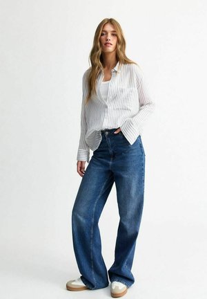 Vit och grå randig skjorta med knappar, avslappnad passform, i kombination med högmidjade blå jeans. Modellen bär vita sneakers med bruna detaljer.