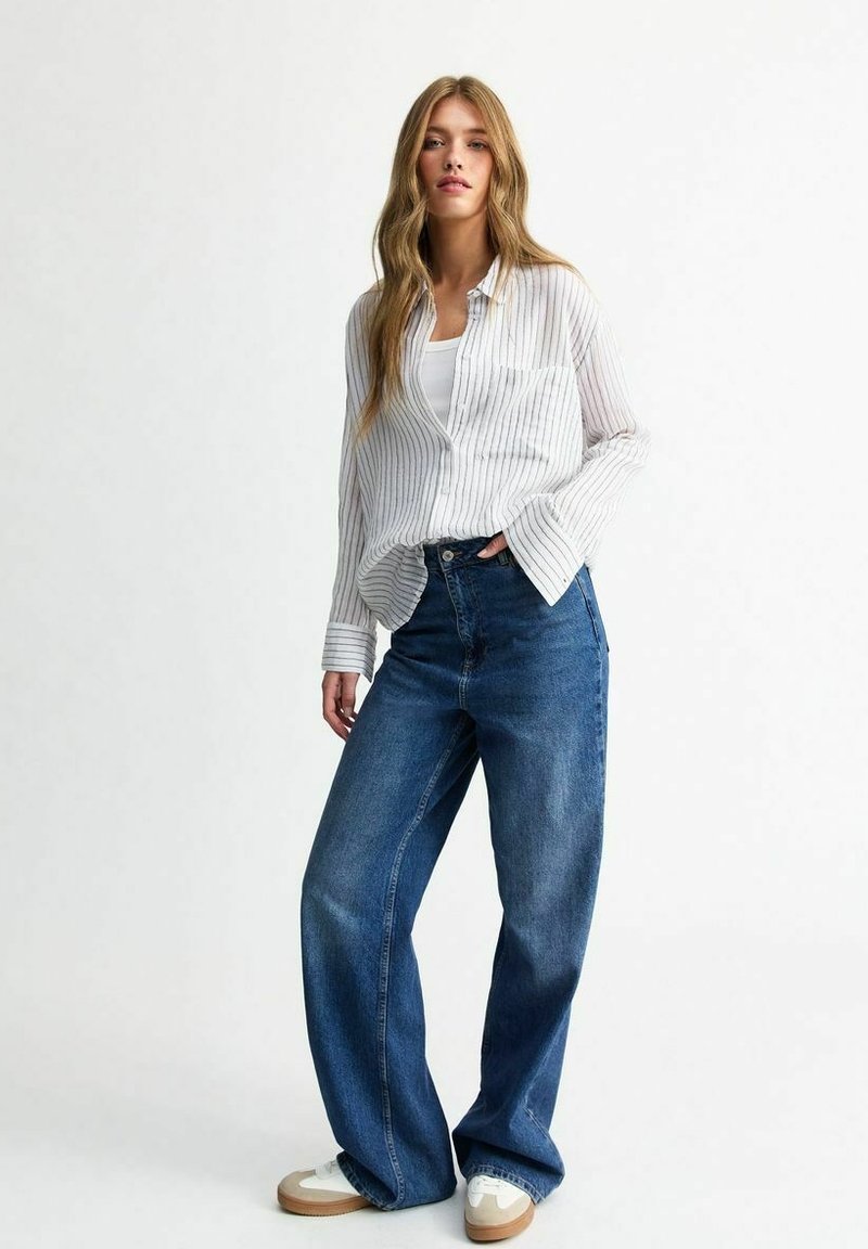 Vit och grå randig skjorta med knappar, avslappnad passform, i kombination med högmidjade blå jeans. Modellen bär vita sneakers med bruna detaljer.
