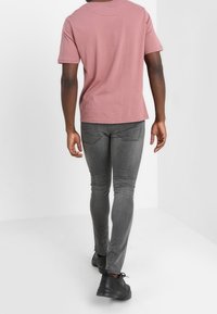 Rosa bomullströja, figurnära design, grå skinny jeans med bakfickor och svarta sneakers. Enkel, avslappnad outfit mot en vit bakgrund.