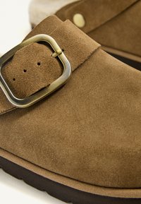 Hnedá semišová topánka s veľkou mosadznou prackou, zaoblenou špičkou a hrubou čiernou podrážkou.