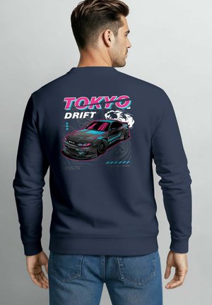 Man wearing navy sweatshirt with colorful graphic of drifting sports car and text "Tokyo Drift" on the back.