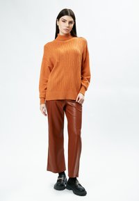 Maglione arancione a collo alto lavorato a maglia con un motivo a foglie testurizzate, abbinato a pantaloni a gamba larga in ecopelle marrone e mocassini neri spessi.