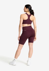 Kvinna i vinröda träningsshorts och sport-bh, stående och tittande åt sidan, med vita sneakers och vita strumpor.