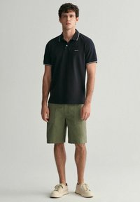 Čierne polo tričko s bielym lemovaním, skombinované s olivovými šortkami. Ľahký materiál, voľný strih. Model má na sebe béžové tenisky.