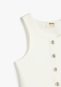 Vestido blanco sin mangas con cuello redondo, presenta botones metálicos en la parte delantera y una textura suave. La etiqueta dice "Koton Kids".