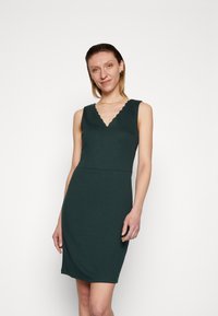 Vestido sem mangas de um verde escuro, com decote em V em forma de ondas, silhueta ajustada e textura suave, adequado para ocasiões casuais ou formais.