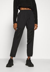 Pantalones deportivos negros con paneles de malla, cintura elástica y puños elásticos, combinados con zapatillas negras. Sin gráficos ni patrones visibles.