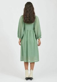 Zelené středně dlouhé šaty s texturovanou tkaninou, nabíranými rukávy, přiléhavým pasem a splývavou sukní; nošeny s bílými kotníkovými botami.