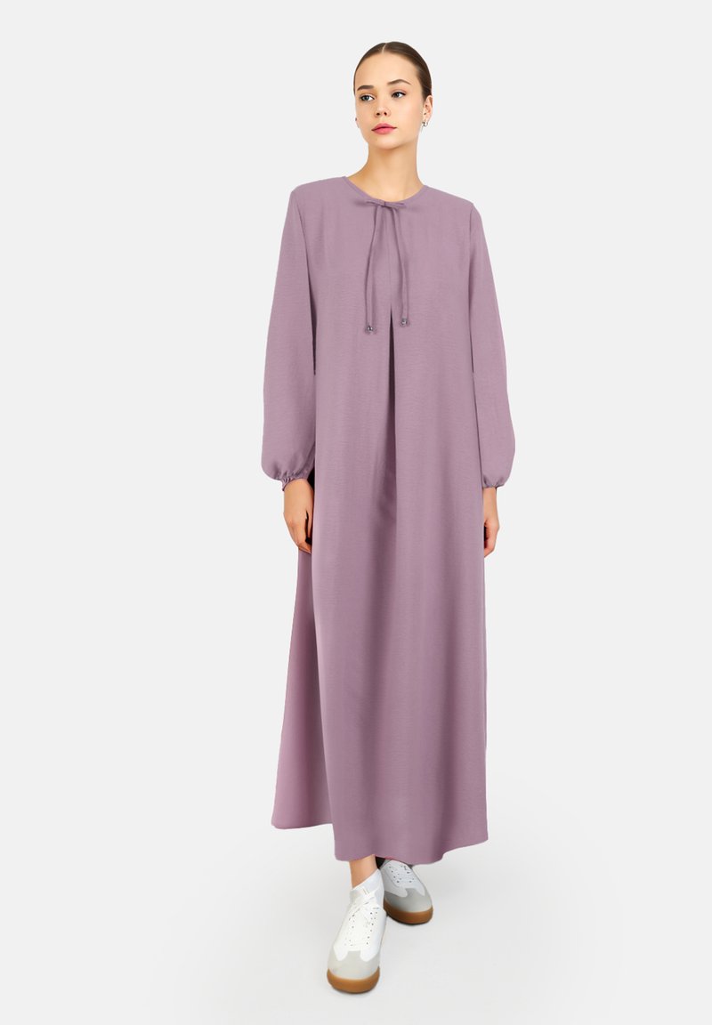 Langes, lila Kleid mit langen Ärmeln, elastischen Manschetten und einem gebundenen Ausschnitt. Getragen mit weißen Sneakers mit brauner Sohle.