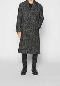 Černobílý dvouřadý kabát ve vzoru rybí kosti z husté látky, s dlouhým střihem a bočními kapsami, nošený přes černé kalhoty a boty.