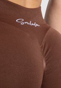 Bruine high-waisted leggings van rekbare stof, met een gladde textuur en een wit logo nabij de tailleband.